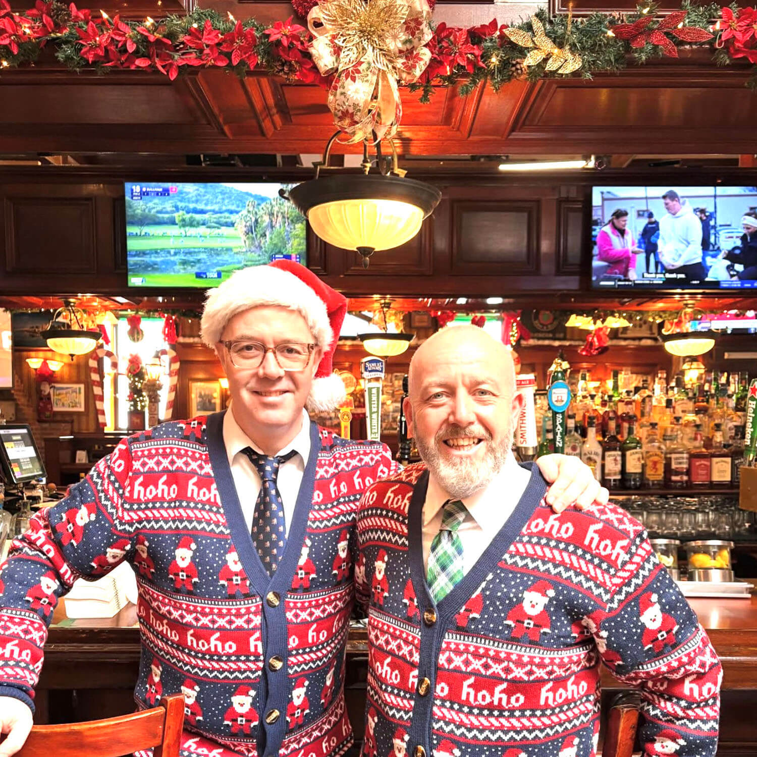 Two men smiling behind a bar wearing matching festive Christmas sweaters with Santa and 'ho ho ho' patterns, one man in a Santa hat.