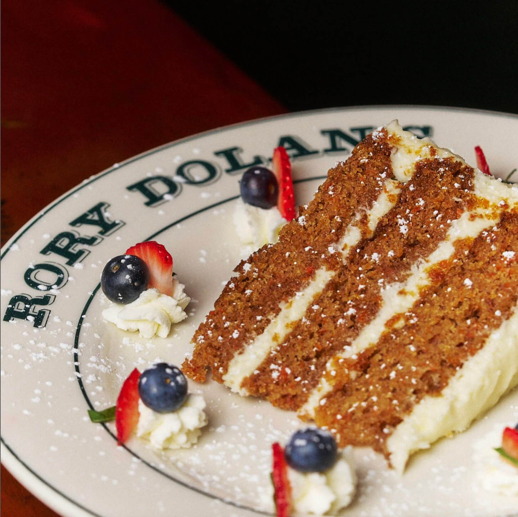Slice of three-layer carrot cake with cream cheese frosting, garnished with blueberries, strawberry slices, and whipped cream on a Róry Dolan’s plate.