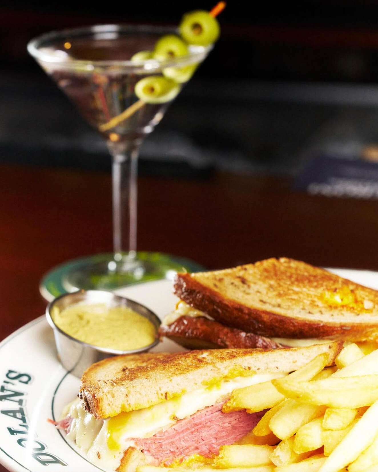 Corned beef sandwich with melted cheese and French fries on a restaurant plate with mustard and a martini glass garnished with green olives in the background.