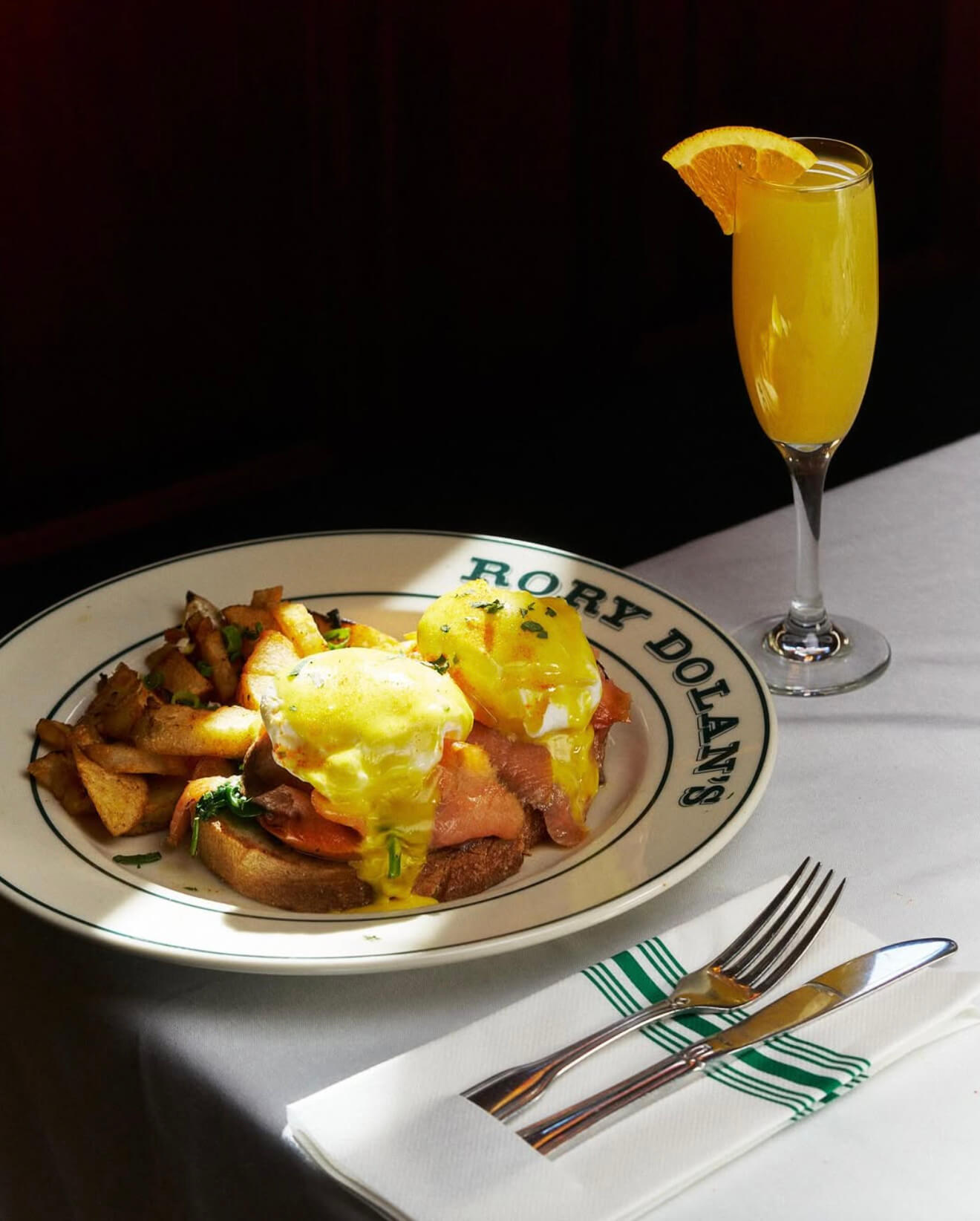 Eggs Benedict with smoked salmon, home fries on a Rory Dolan's plate, next to a mimosa glass with an orange slice on a white tablecloth.