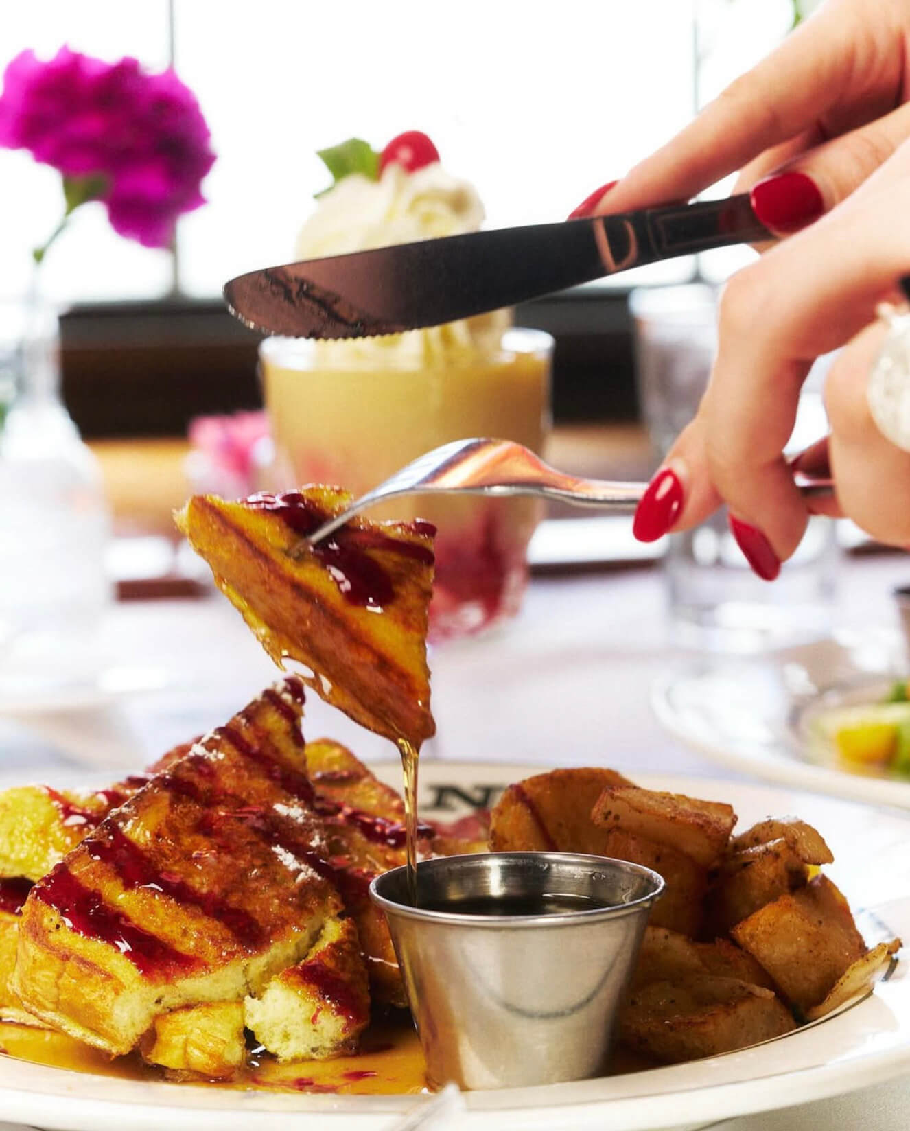 Close-up of French toast drizzled with syrup and berry sauce, held by fork and knife with a side of roasted potatoes.