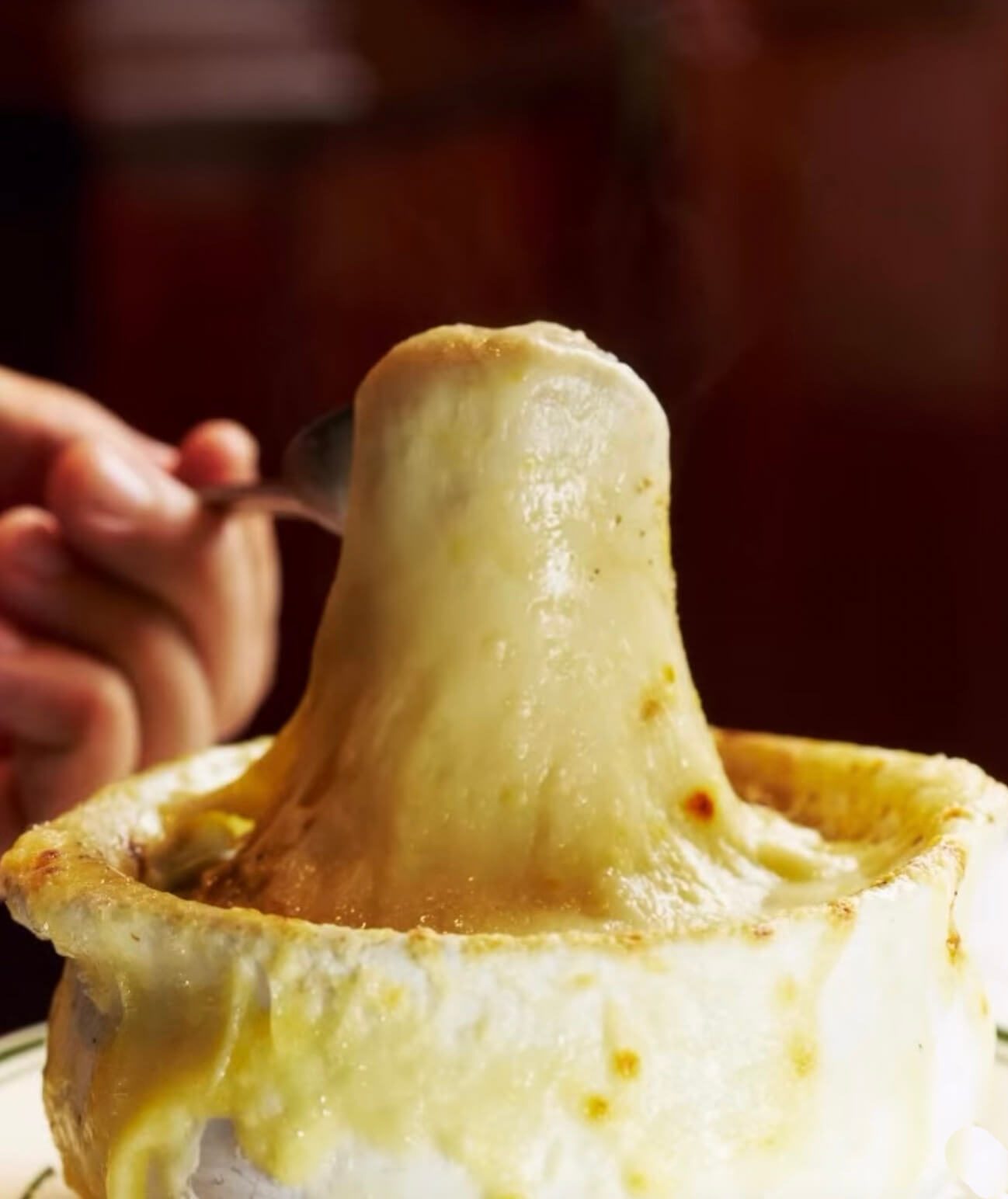 Hand lifting a spoonful of melted cheese from a bowl of French onion soup with melted cheese overflow.