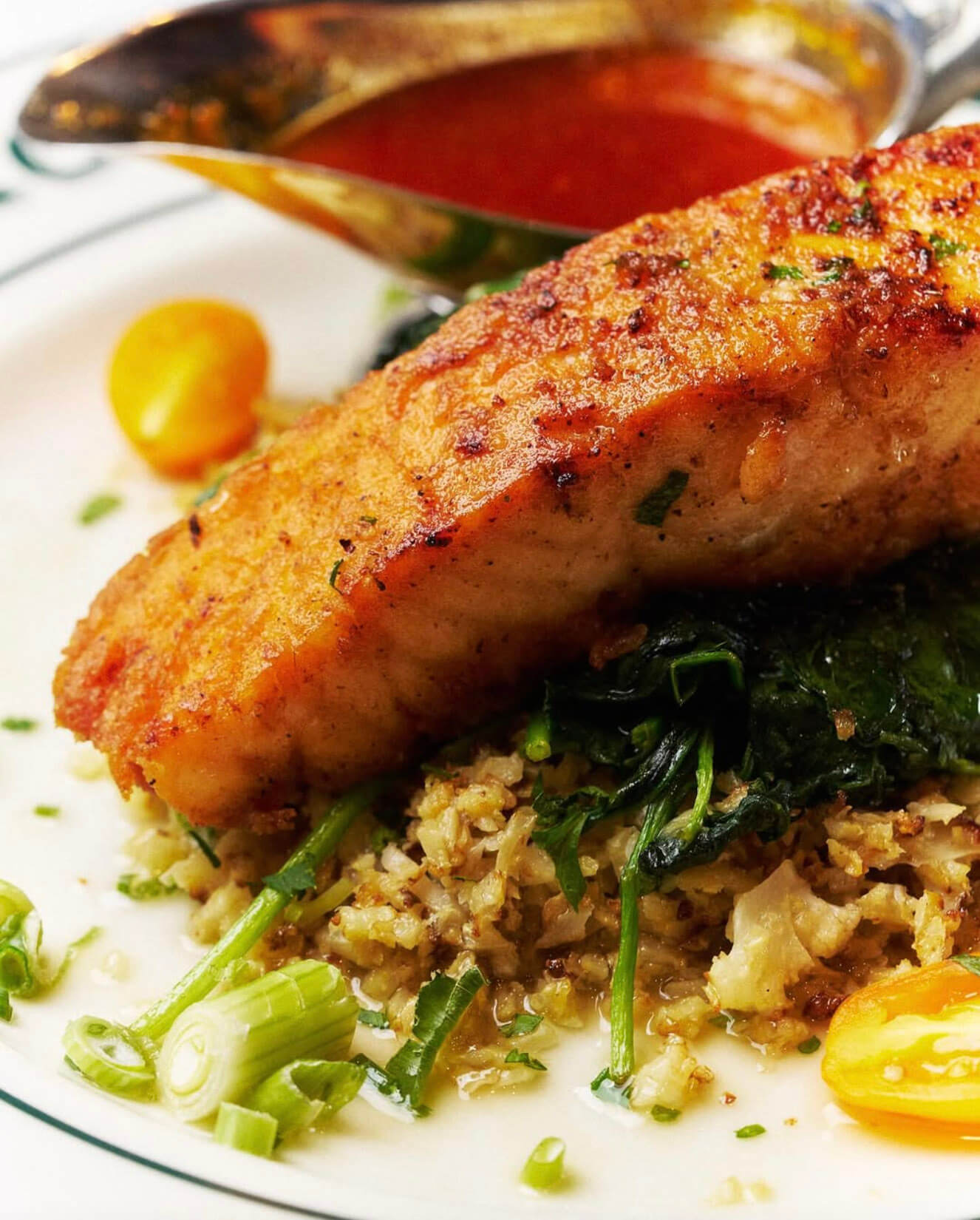 Close-up of a grilled salmon fillet on sautéed spinach and grain, garnished with green onions and yellow cherry tomatoes, with a sauce boat in the background.
