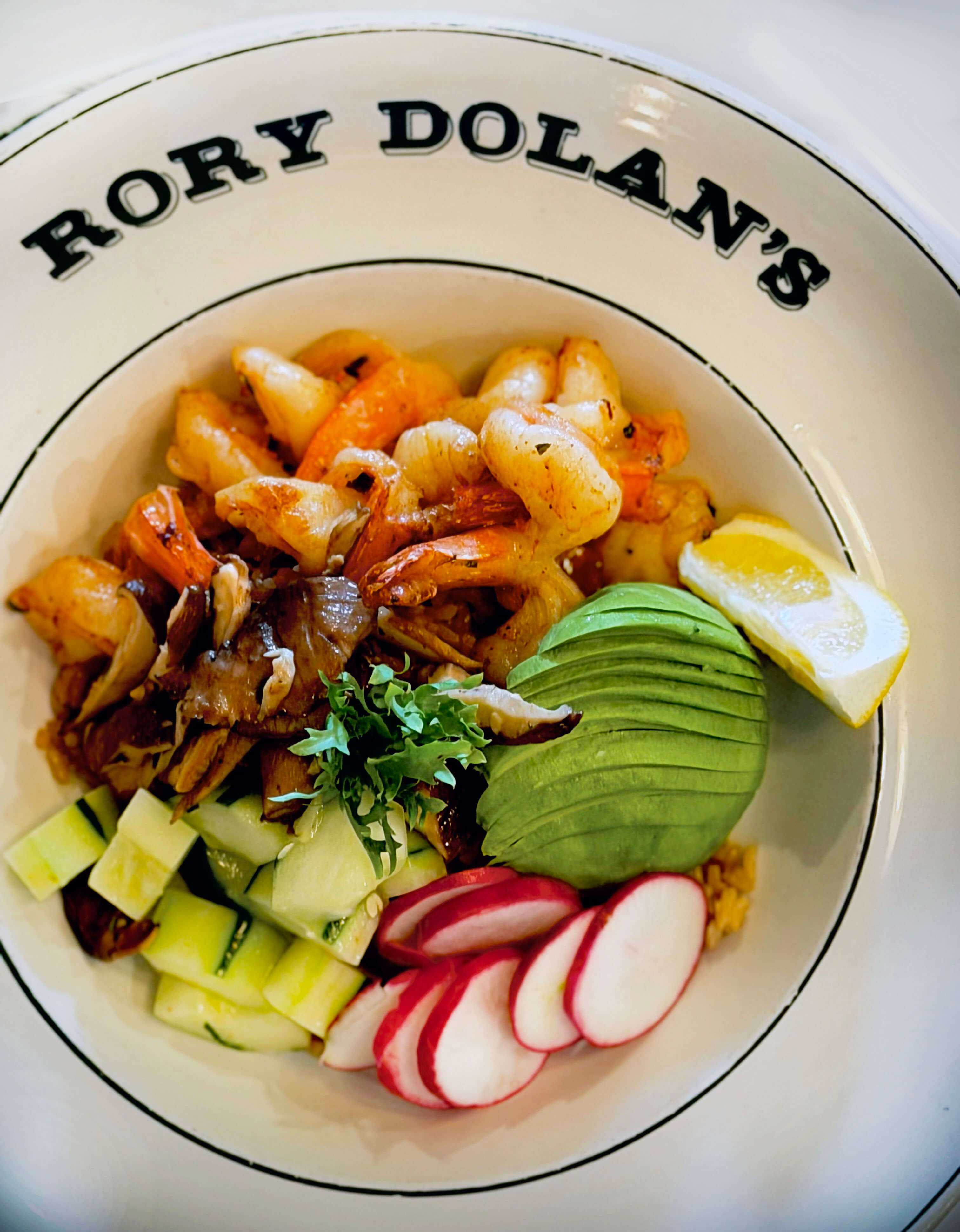 Bowl with grilled shrimp, sliced avocado, radishes, cucumber, greens, and lemon wedge.