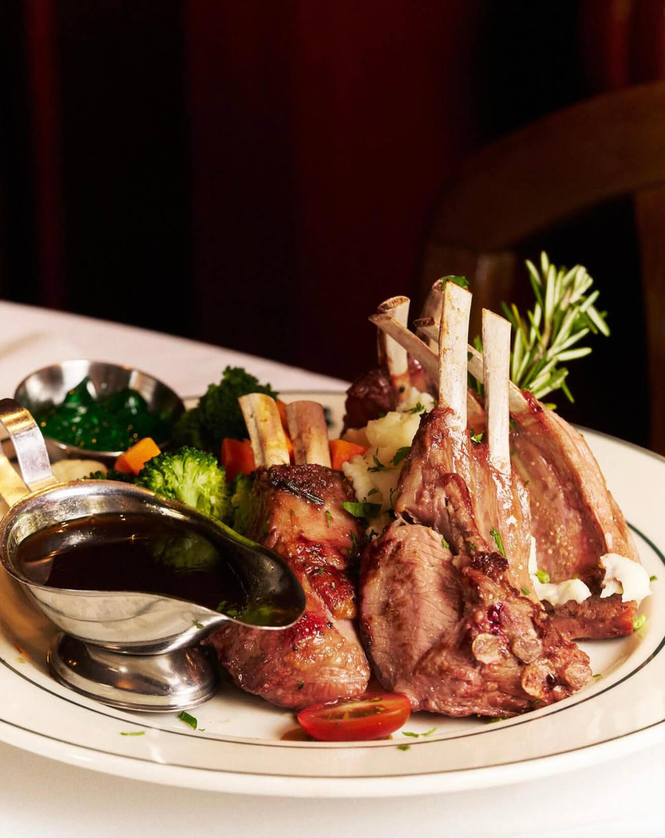 Plate of grilled lamb chops garnished with rosemary, served with mixed steamed vegetables, mashed potatoes, and a side of dark brown gravy sauce.