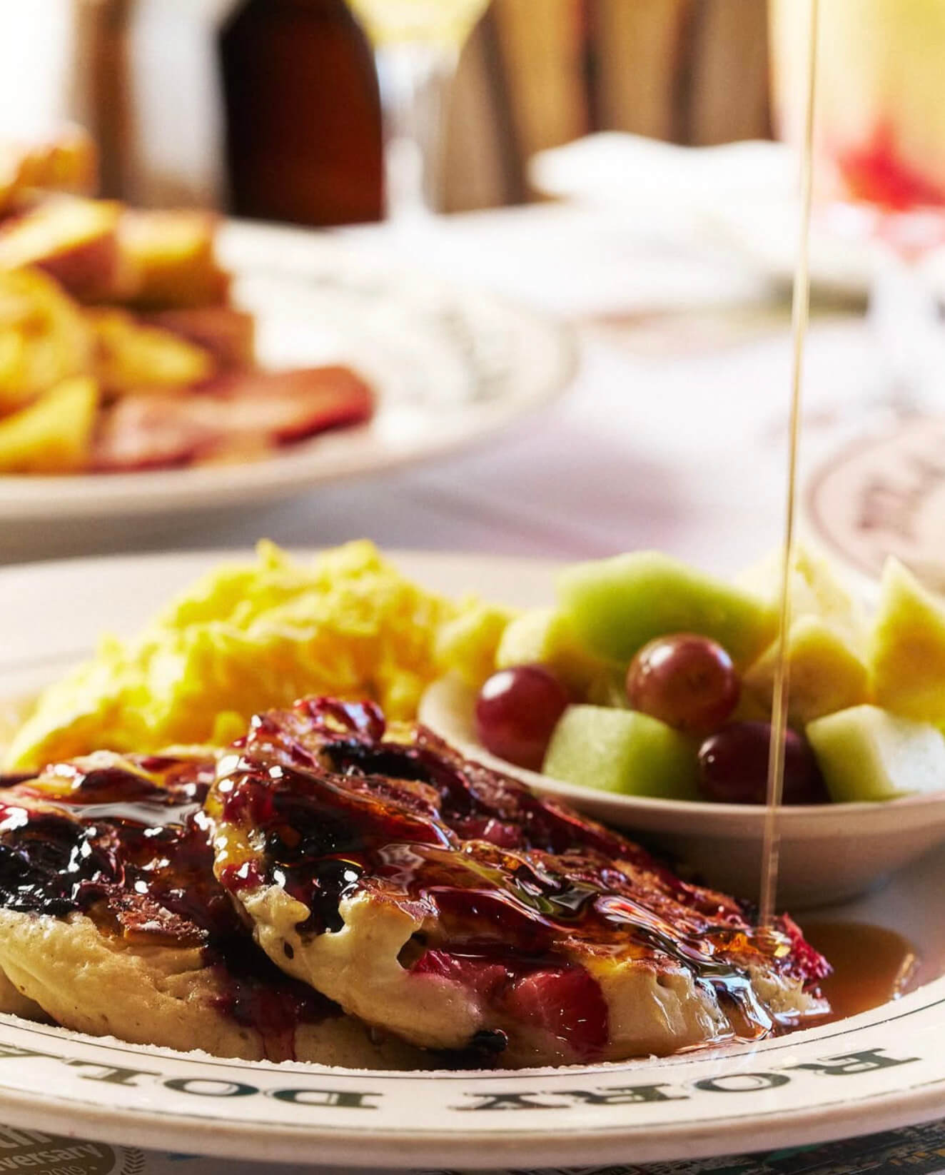 Stack of berry pancakes drizzled with syrup, scrambled eggs, and a bowl of mixed fruit including grapes and melon on a plate.