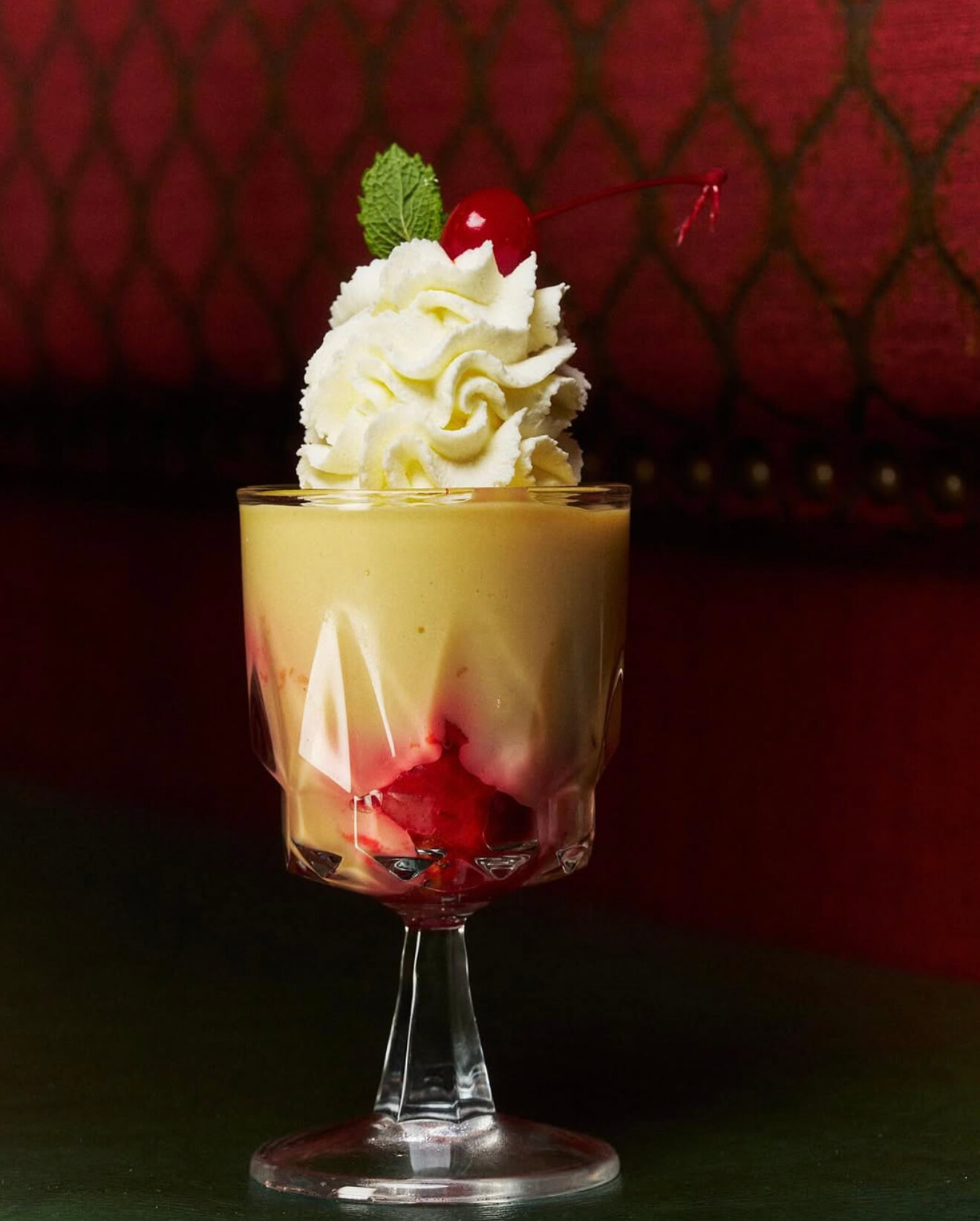 Glass dessert with custard, red fruit syrup, whipped cream, a cherry, and a mint leaf on top.