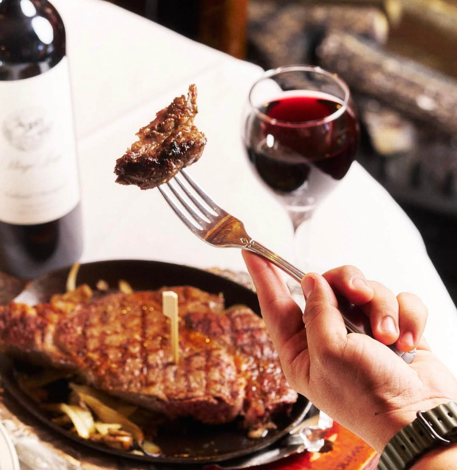 Hand holding a fork with a piece of grilled steak, a full steak on a plate, and a glass of red wine on a white tablecloth.