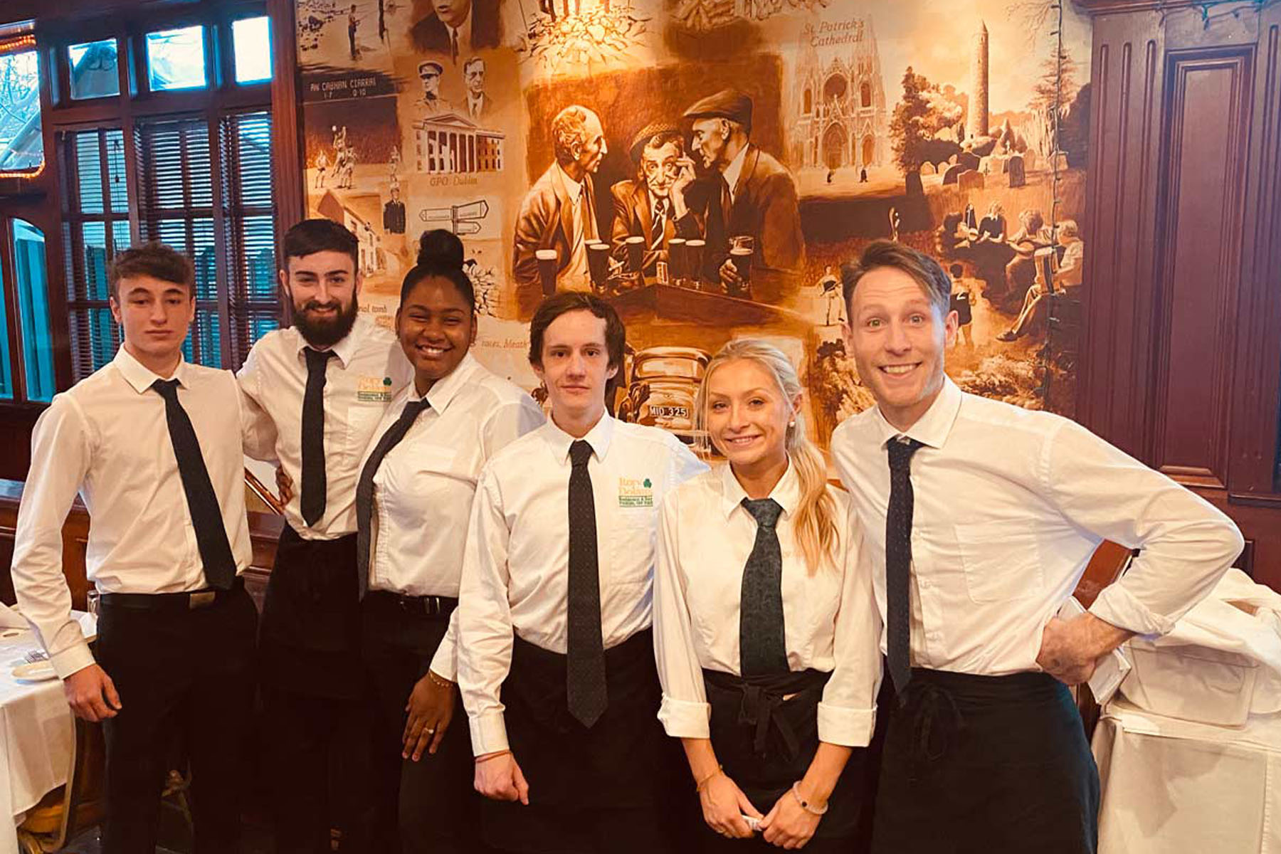 Group of six smiling restaurant staff in white shirts, black ties, and aprons posing indoors in front of a mural.